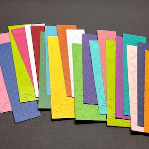 May include: A collection of patterned cardstock in various colors, including pink, green, blue, yellow, orange, and purple. The cardstock is arranged in a fan shape, with the edges of each sheet visible.