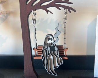 May include: A wooden Halloween decoration featuring a ghost on a swing. The ghost is holding a mug, with steam rising. The swing is suspended from a tree-shaped structure. The color palette includes brown, black, and white.