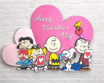 Peut inclure: Une pancarte en forme de cœur rose avec le texte "Happy Valentine's Day" et un groupe de personnages de Peanuts tenant des cartes de la Saint-Valentin. Les personnages incluent Charlie Brown, Snoopy, Lucy, Linus et Sally.