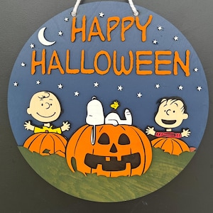 Op de afbeelding: Ronde houten Halloween-decoratie met Charlie Brown, Snoopy en Linus op een pompoen. De woorden "HAPPY HALLOWEEN" staan in oranje op een blauwe achtergrond met sterren en een maan. Een wit touwtje is bevestigd om op te hangen.