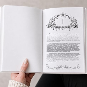 Könnte beinhalten: Ein offenes Buch mit einem dekorativen schwarz-weißen Design auf der rechten Seite. Die Seite zeigt die Zahl "1" in einem detaillierten Rahmen mit Text darunter. Eine Hand mit einem Ring hält das Buch.