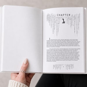 Könnte beinhalten: Ein offenes Buch mit weißen Seiten. Die rechte Seite zeigt das Wort "KAPITEL" über der Zahl "1" und ein vertikales Binärcode-Design. Die linke Seite ist leer. Das Buch wird von einer Hand offen gehalten.