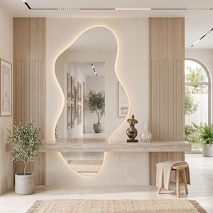 Op de afbeelding: Een eigentijds interieurontwerp met een grote, organisch gevormde spiegel met geïntegreerde verlichting. De ruimte omvat houten panelen, een marmeren plank, decoratieve items en potplanten, wat een serene en stijlvolle sfeer creëert.