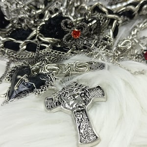 Puede incluir: Un surtido de collares y colgantes plateados sobre una superficie blanca y esponjosa. La colecci&oacute;n incluye colgantes de cruz, dijes en forma de coraz&oacute;n con detalles rojos y un colgante en forma de coraz&oacute;n negro. La joyer&iacute;a es intrincadamente detallada.