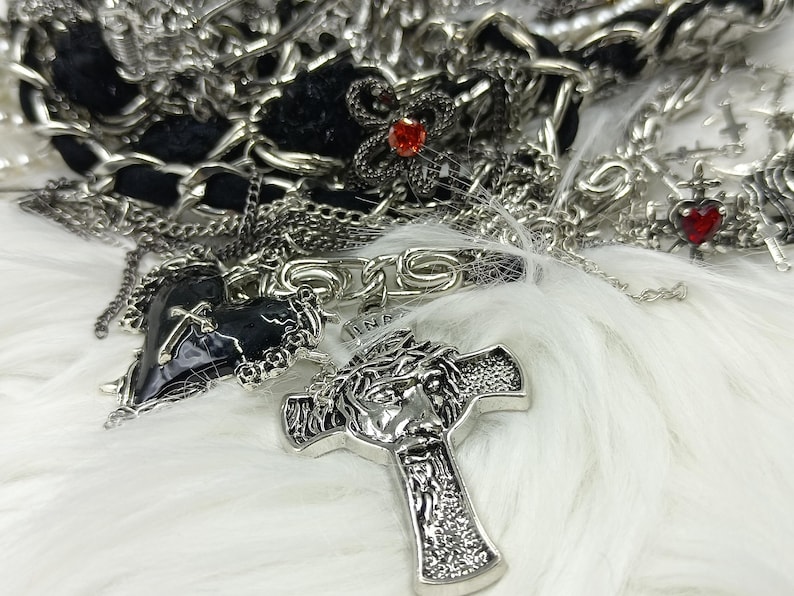 Puede incluir: Un surtido de collares y colgantes plateados sobre una superficie blanca y esponjosa. La colecci&oacute;n incluye colgantes de cruz, dijes en forma de coraz&oacute;n con detalles rojos y un colgante en forma de coraz&oacute;n negro. La joyer&iacute;a es intrincadamente detallada.