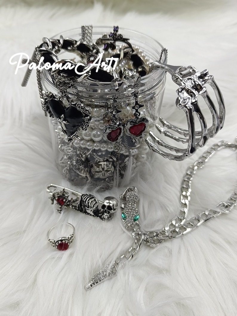 Puede incluir: Una colecci&oacute;n de joyas g&oacute;ticas, que incluye anillos, pendientes, pulseras y un collar. Las joyas presentan dise&ntilde;os de coraz&oacute;n, calavera y mano de esqueleto, con detalles en rojo y negro. Los art&iacute;culos se exhiben en un tarro transparente y sobre una superficie blanca y esponjosa. El texto "Paloma Art" es visible.