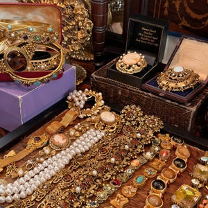 May include: A collection of vintage gold-toned jewelry, including bracelets, brooches, and necklaces, displayed on a wooden surface. The jewelry features pearls, gemstones, and intricate designs. Some pieces are in velvet-lined boxes.