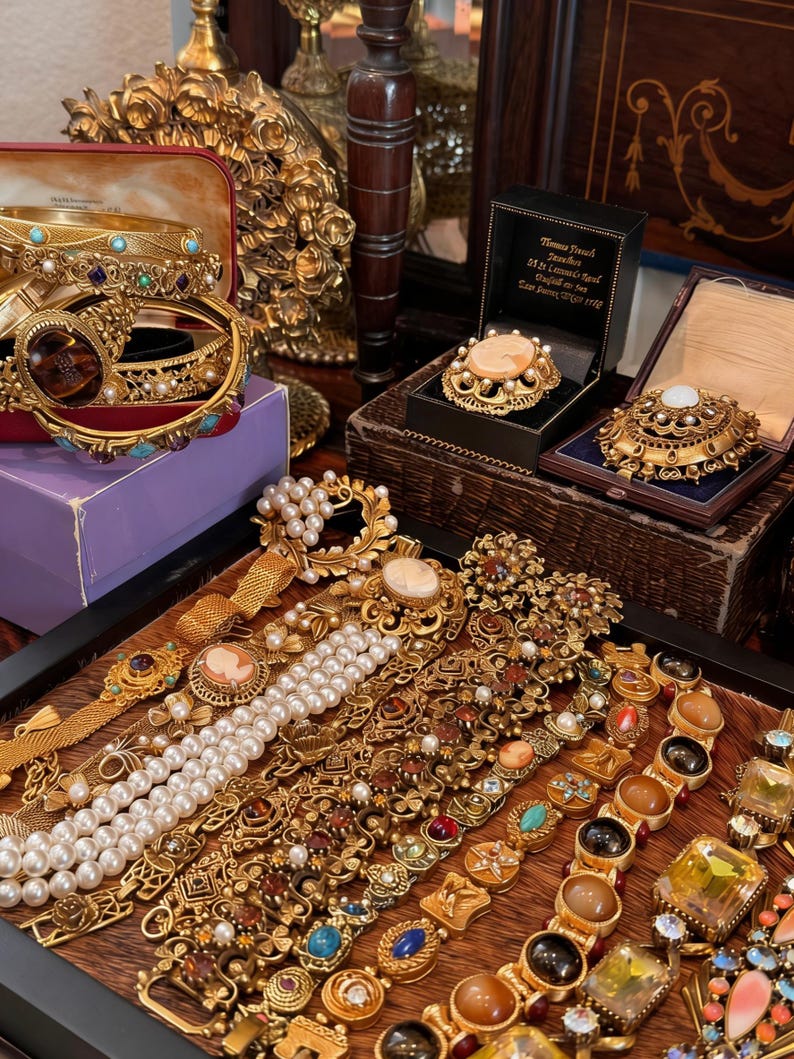 May include: A collection of vintage gold-toned jewelry, including bracelets, brooches, and necklaces, displayed on a wooden surface. The jewelry features pearls, gemstones, and intricate designs. Some pieces are in velvet-lined boxes.