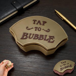 May include: A beige, fan-shaped box with the text "TAP TO BUBBLE" in brown. A smaller, matching piece is next to it, and a hand holds a small, round piece. A black notebook and gold pen are also visible.