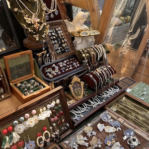 May include: A display of vintage jewelry, including necklaces, earrings, rings, and bracelets. The jewelry is arranged on velvet-lined trays and stands, showcasing pearls, gemstones, and ornate designs. A small statue of an angel is also present.