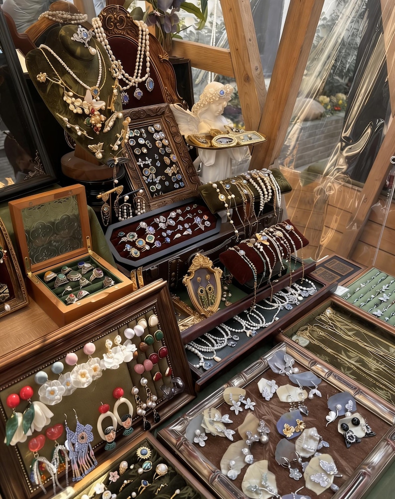 May include: A display of vintage jewelry, including necklaces, earrings, rings, and bracelets. The jewelry is arranged on velvet-lined trays and stands, showcasing pearls, gemstones, and ornate designs. A small statue of an angel is also present.
