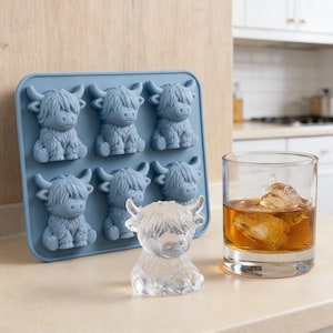 Puede incluir: Una bandeja de cubitos de hielo de silicona azul con seis moldes en forma de vaca de las Tierras Altas. Un cubito de hielo transparente con forma de vaca se encuentra junto a la bandeja, y un vaso de líquido ámbar con un cubito de hielo con forma de vaca está a la derecha. Utensilios de cocina y bar.