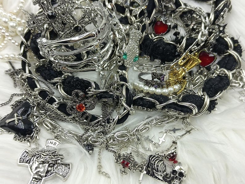 Puede incluir: Un surtido de joyas plateadas, que incluyen collares, pulseras y anillos, con detalles en negro y adornos en forma de coraz&oacute;n rojo. La colecci&oacute;n presenta cruces, calaveras y hileras de perlas, creando una est&eacute;tica g&oacute;tica o punk.