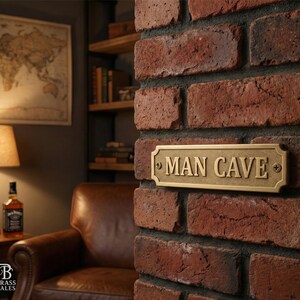 May include: A rectangular brass sign with the words "MAN CAVE" in capital letters is mounted on a red brick wall. The sign has a decorative border and is secured with screws. The background includes a map, a lamp, and a leather armchair.