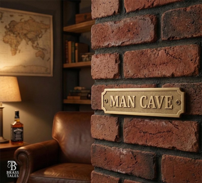 May include: A rectangular brass sign with the words "MAN CAVE" in capital letters is mounted on a red brick wall. The sign has a decorative border and is secured with screws. The background includes a map, a lamp, and a leather armchair.