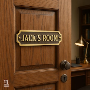 May include: A rectangular brass plaque with the text "JACK'S ROOM" in gold lettering is mounted on a wooden door. The plaque has a dark brown background and a decorative border. The door is open, revealing a desk and lamp.