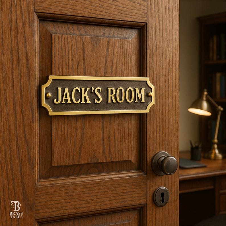 May include: A rectangular brass plaque with the text "JACK'S ROOM" in gold lettering is mounted on a wooden door. The plaque has a dark brown background and a decorative border. The door is open, revealing a desk and lamp.