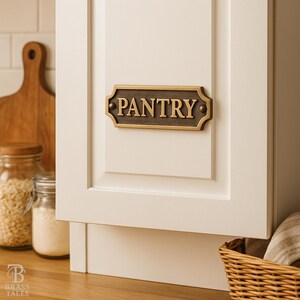 May include: A bronze-colored pantry sign with the word "PANTRY" in capital letters is mounted on a white cabinet door. The sign has a decorative border and is part of a kitchen organization theme.