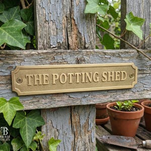 May include: A rectangular brass sign that reads "THE POTTING SHED" is mounted on a weathered wooden fence. The sign is secured with two screws and has decorative edges. Green ivy and small terracotta pots are in the background.