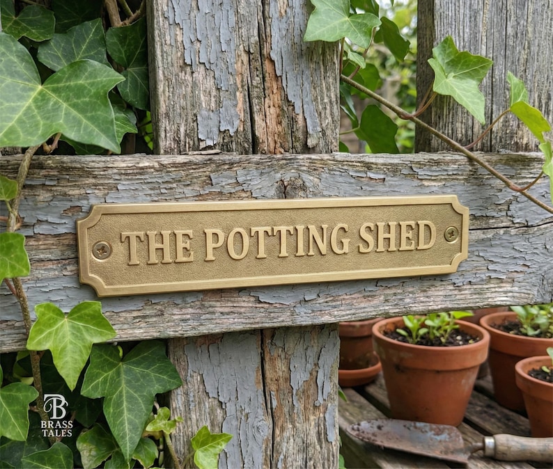 May include: A rectangular brass sign that reads "THE POTTING SHED" is mounted on a weathered wooden fence. The sign is secured with two screws and has decorative edges. Green ivy and small terracotta pots are in the background.