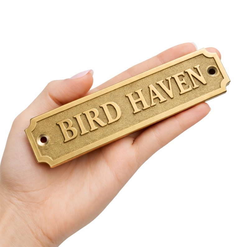 May include: A rectangular, gold-colored metal sign with the words "BIRD HAVEN" in raised lettering. The sign has rounded corners and two holes for mounting. The sign is held in a hand.
