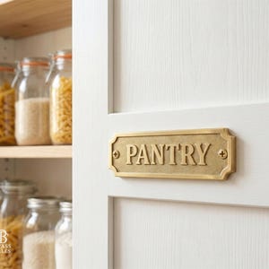 May include: A rectangular brass pantry sign with the word "PANTRY" in bold, raised letters is mounted on a white door. The sign has decorative edges and is secured with small screws. Jars of pasta and rice are visible on a shelf.