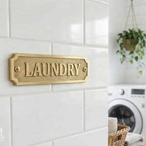 May include: A rectangular brass laundry sign with the word "LAUNDRY" in raised letters. The sign is mounted on white tile and has decorative edges. The sign is attached with two screws, one on each side.