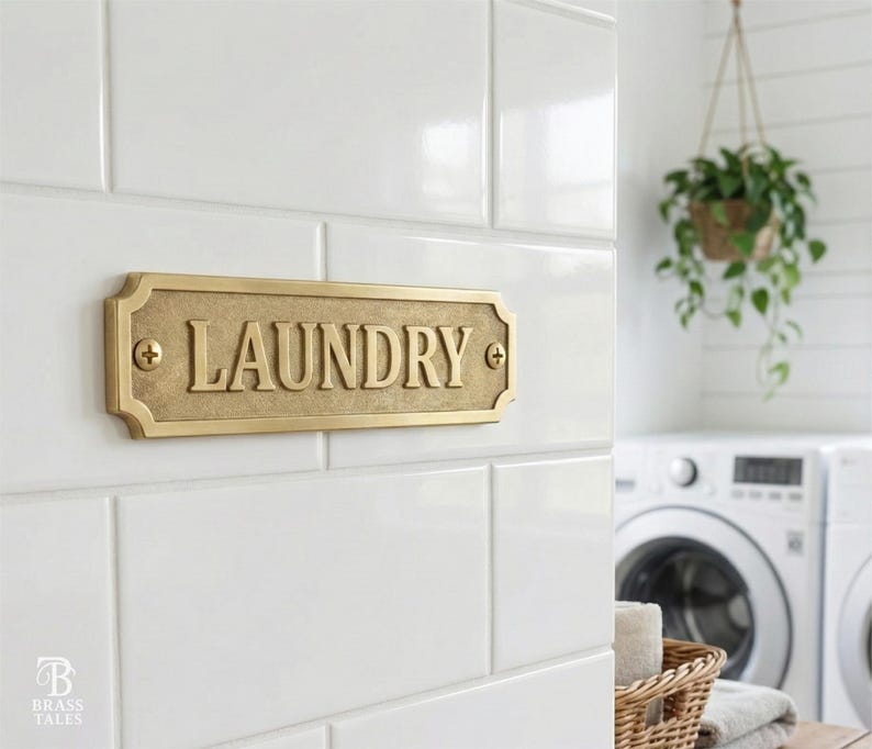 May include: A rectangular brass laundry sign with the word "LAUNDRY" in raised letters. The sign is mounted on white tile and has decorative edges. The sign is attached with two screws, one on each side.