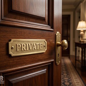 May include: A close-up of a wooden door with a brass "PRIVATE" sign and matching door handle. The sign is rectangular with rounded corners and is mounted on the door with two screws. The door is open, revealing a hallway.