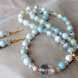 Op de afbeelding: Een set lichtblauwe, witte en gouden kralen sieraden met een grote kristallen kraal. De set bevat een ketting, een armband en een paar oorbellen.