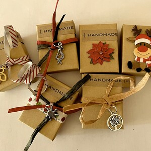 Puede incluir: Varias cajas de regalo peque&ntilde;as, marrones, atadas con cintas y adornadas con abalorios. Algunas cajas tienen la palabra "HANDMADE" impresa. Los abalorios incluyen una flor, una figura de Kokopelli, un sol y un &aacute;rbol de la vida. Una caja est&aacute; decorada con una flor de Pascua y otra con un reno.
