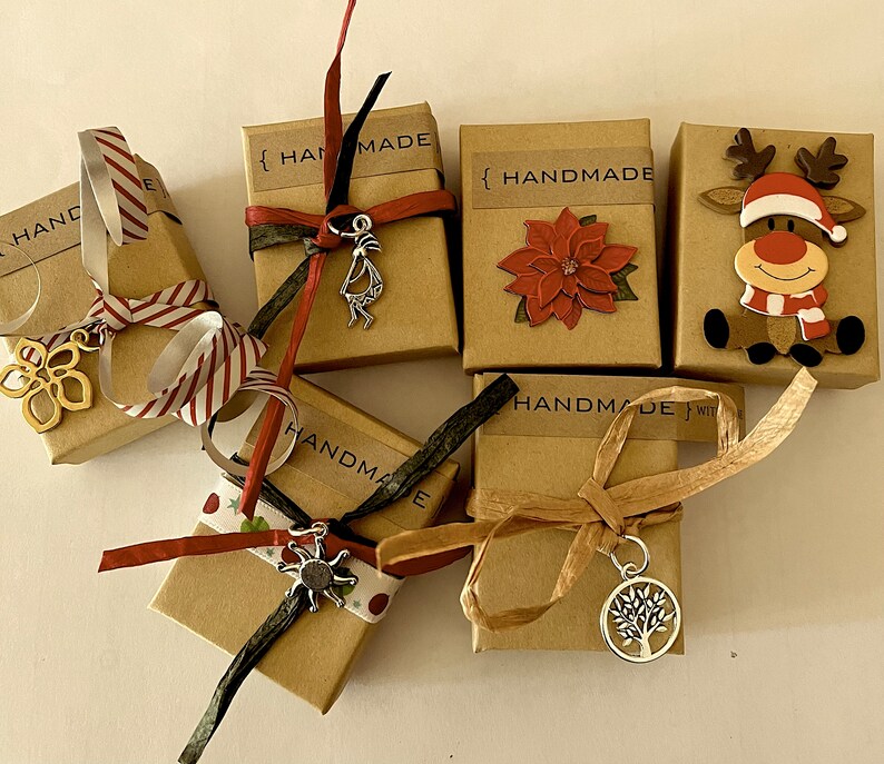 Puede incluir: Varias cajas de regalo peque&ntilde;as, marrones, atadas con cintas y adornadas con abalorios. Algunas cajas tienen la palabra "HANDMADE" impresa. Los abalorios incluyen una flor, una figura de Kokopelli, un sol y un &aacute;rbol de la vida. Una caja est&aacute; decorada con una flor de Pascua y otra con un reno.