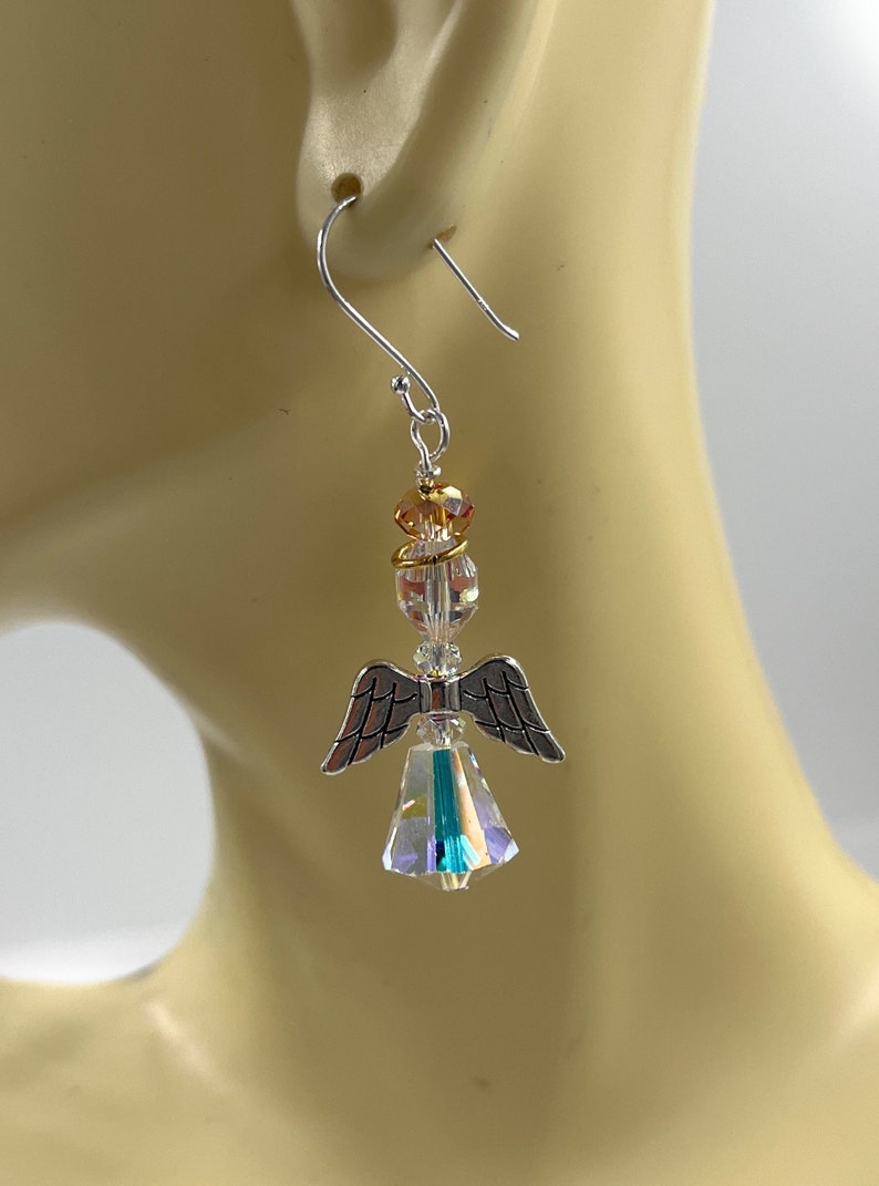 Puede incluir: Un solo pendiente de &aacute;ngel plateado con cuerpo y alas de cristal transparente e iridiscente. El &aacute;ngel tiene un halo y una cabeza dorados. El pendiente cuelga de un gancho plateado, mostrando un dise&ntilde;o delicado y artesanal.