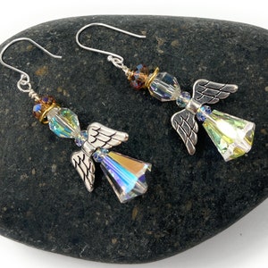 Puede incluir: Un par de pendientes con forma de &aacute;ngel con alas plateadas y cuerpos de cristal iridiscente. Los pendientes tienen cierre de gancho y se exhiben sobre una piedra gris oscura. Los cristales reflejan una gama de colores.