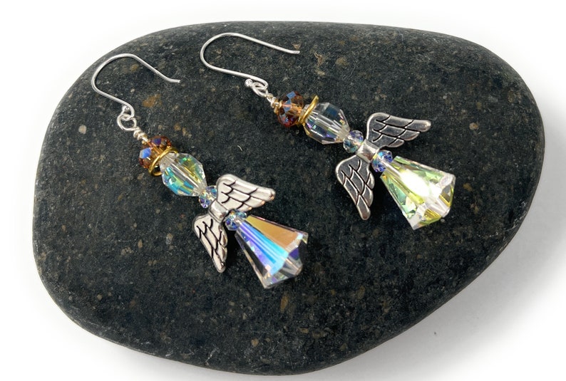 Puede incluir: Un par de pendientes con forma de &aacute;ngel con alas plateadas y cuerpos de cristal iridiscente. Los pendientes tienen cierre de gancho y se exhiben sobre una piedra gris oscura. Los cristales reflejan una gama de colores.