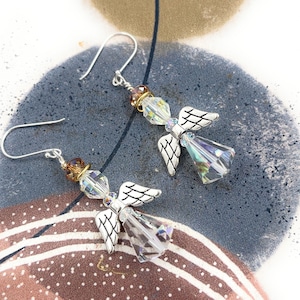 Puede incluir: Un par de pendientes de &aacute;ngel con alas plateadas y cuerpos de cristal transparente facetado. Los pendientes tienen ganchos plateados y est&aacute;n adornados con cuentas marrones y doradas. Se exhiben sobre una superficie estampada azul y marr&oacute;n.