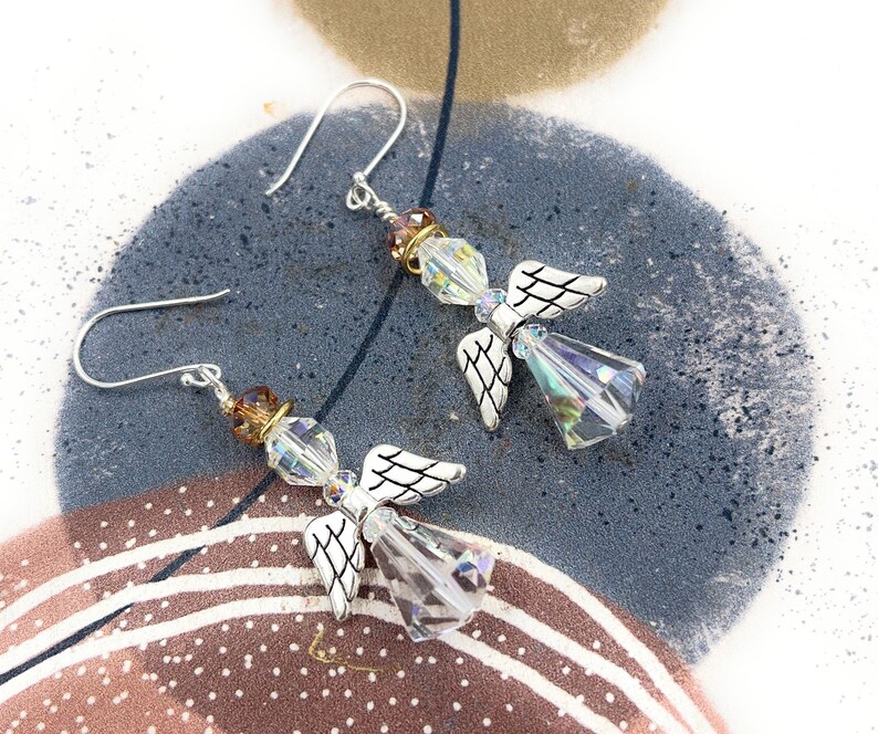 Puede incluir: Un par de pendientes de &aacute;ngel con alas plateadas y cuerpos de cristal transparente facetado. Los pendientes tienen ganchos plateados y est&aacute;n adornados con cuentas marrones y doradas. Se exhiben sobre una superficie estampada azul y marr&oacute;n.