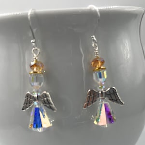 Puede incluir: Un par de pendientes de &aacute;ngel con alas plateadas y cuerpos de cristal iridiscente. Los pendientes tienen un gancho plateado y est&aacute;n acentuados con cuentas de color &aacute;mbar. Los pendientes se muestran sobre un fondo blanco.