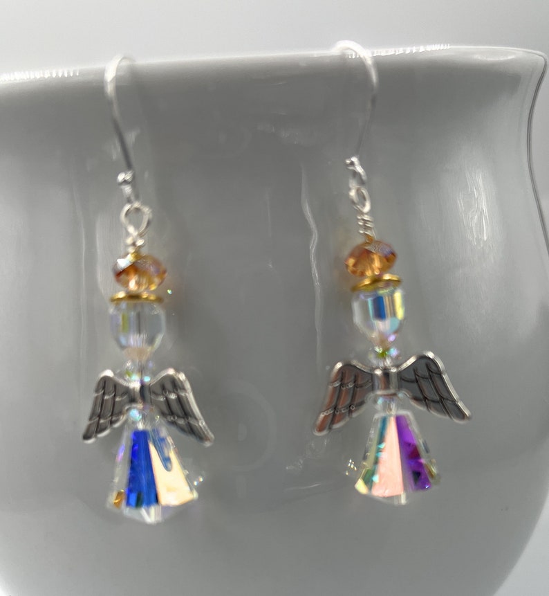 Puede incluir: Un par de pendientes de &aacute;ngel con alas plateadas y cuerpos de cristal iridiscente. Los pendientes tienen un gancho plateado y est&aacute;n acentuados con cuentas de color &aacute;mbar. Los pendientes se muestran sobre un fondo blanco.