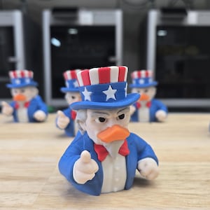May include: A group of Uncle Sam duck figurines. Each figure is dressed in a blue suit, a red bow tie, and a patriotic hat with red and white stripes and stars. The ducks have orange beaks and are giving a thumbs-up.
