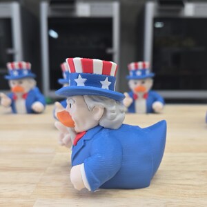 May include: A group of Uncle Sam-themed rubber ducks. Each duck is dressed in a blue suit, a red bow tie, and a star-spangled hat. The ducks are arranged on a light-coloured wooden surface, with a blurred background.