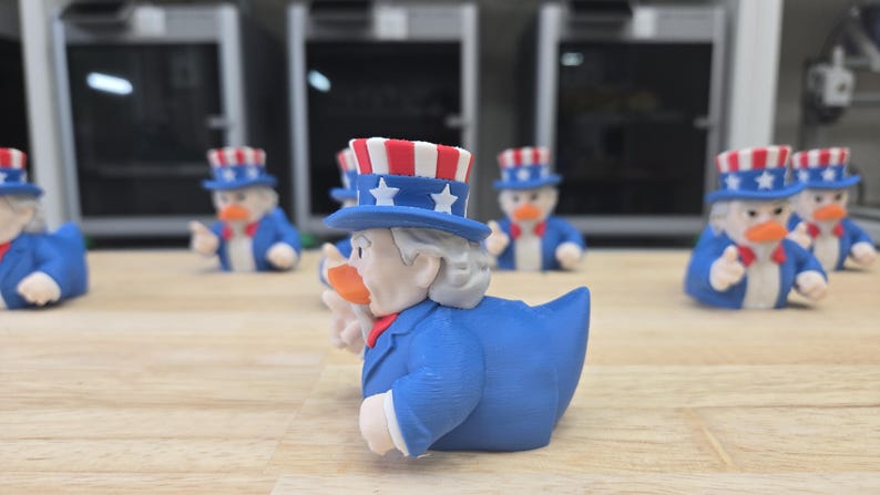 May include: A group of Uncle Sam-themed rubber ducks. Each duck is dressed in a blue suit, a red bow tie, and a star-spangled hat. The ducks are arranged on a light-coloured wooden surface, with a blurred background.