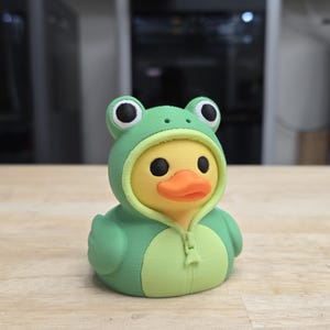 May include: A yellow rubber duck wearing a green frog-themed hooded onesie. The hood features two black and white frog eyes on top, and the onesie includes a molded zipper detail down the center. The duck has a bright orange beak and black eyes.