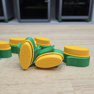 May include: A collection of 3D-printed corn-shaped objects. The objects feature a yellow corn kernel design with green husks and bases. The items are scattered on a light-colored wooden surface.