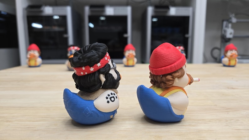 可能包含:两只新奇的鸭子模型。一只鸭子有着黑色毛发,戴着红色头巾,身体呈蓝色。另一只鸭子有着棕色毛发,戴着红色帽子,身体呈蓝色。两只鸭子都摆放在木质桌面上,背景是其他鸭子模型。