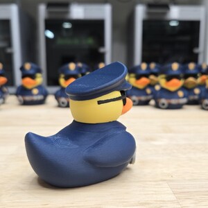 May include: A group of novelty rubber ducks dressed as police officers. The ducks have yellow heads, orange beaks, and wear navy blue uniforms and hats. The ducks are lined up on a wooden surface.