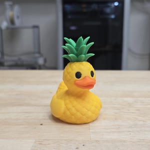 May include: A yellow rubber duck with a pineapple top. The duck has black eyes and an orange beak. The pineapple top is green. The duck is sitting on a wooden surface.
