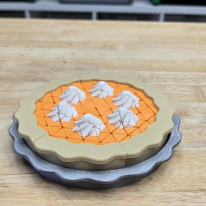 May include: A 3D-printed pie with an orange filling and white whipped cream dollops. The pie sits on a gray plate with a scalloped edge. The crust is tan-colored.