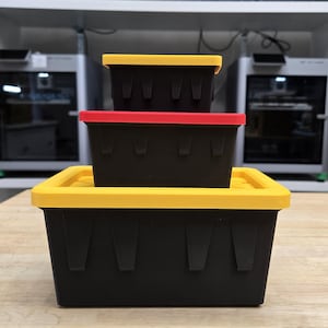 May include: Three black rectangular storage containers with colored lids are stacked on a wooden surface. The lids are yellow and red. The containers have a textured surface and are of varying sizes.