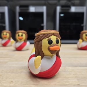 May include: A collection of novelty rubber ducks, each designed to resemble Jesus Christ. The ducks have yellow bodies, brown hair and beards, and wear red and white robes. The ducks are arranged on a wooden surface.